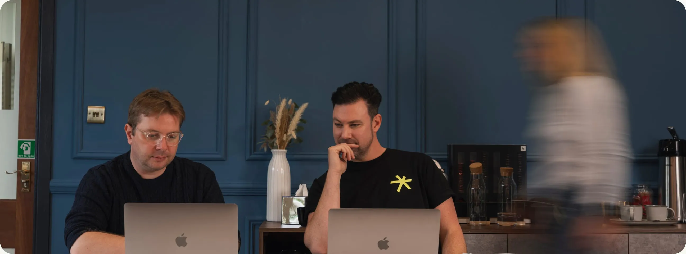 Two developers at their laptops in the studio as a colleague walks past