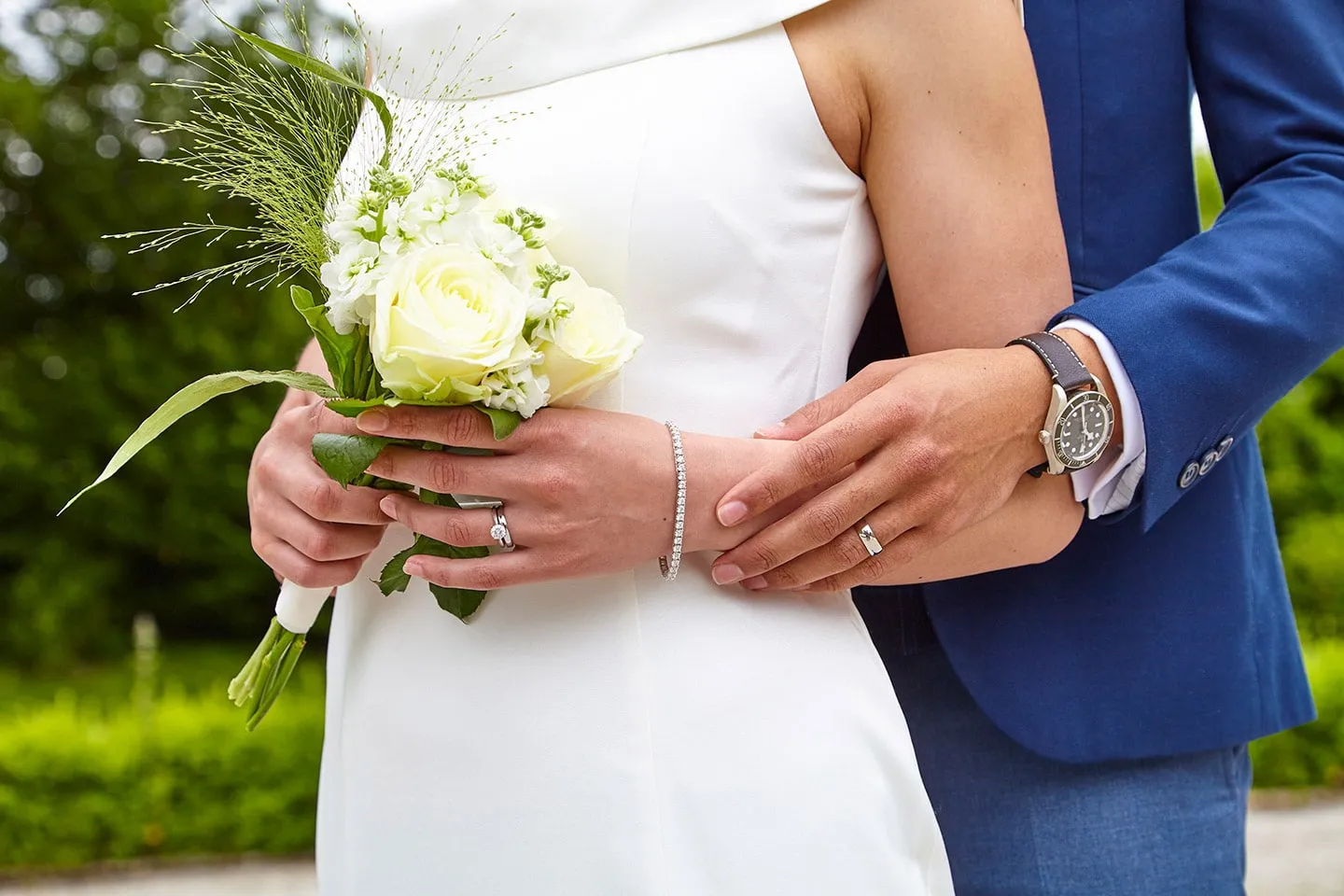 A couple holding hands, showing a Brown & Newirth wedding ring, with a bouquet of white roses