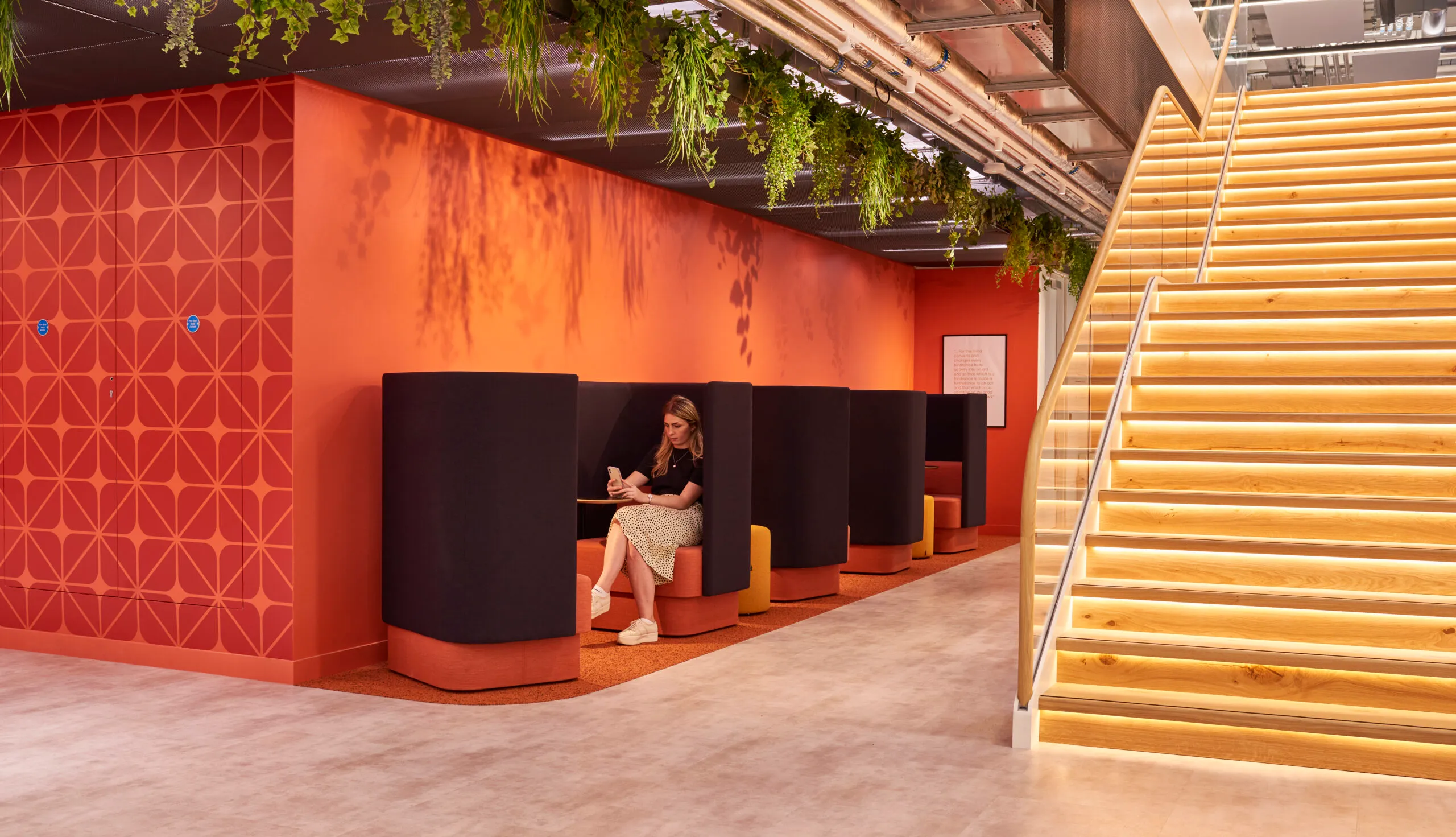 NaughtOne booths along an orange wall, under a staircase and hanging plants