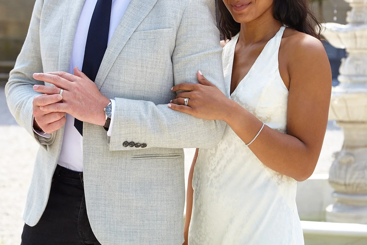 A couple dressed in white, her hand on his arm showing her ring